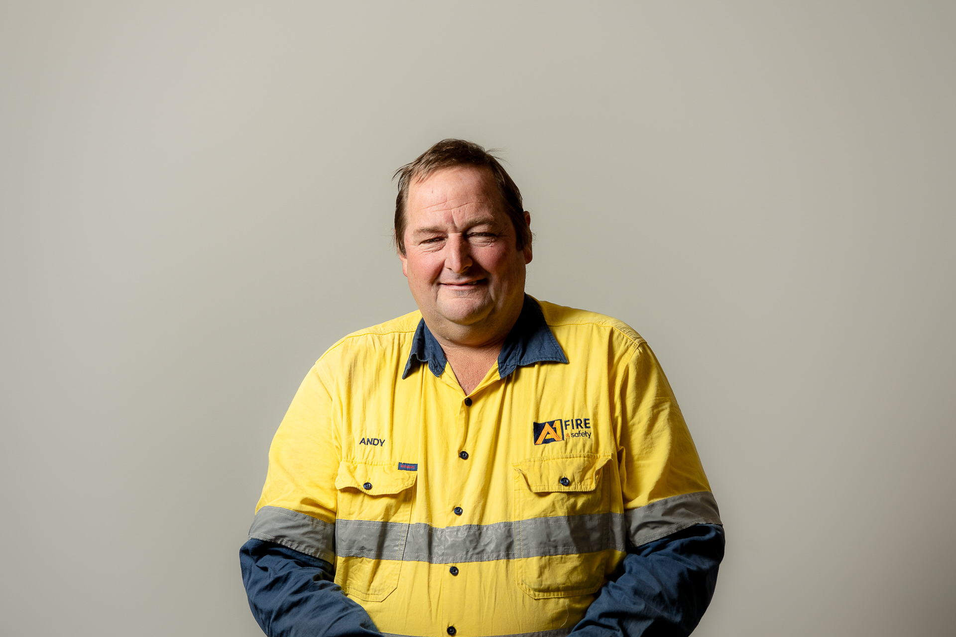 A man wearing a yellow high-visibility work shirt with "Fire Safety" logo stands against a plain gray background, smiling warmly at the camera.