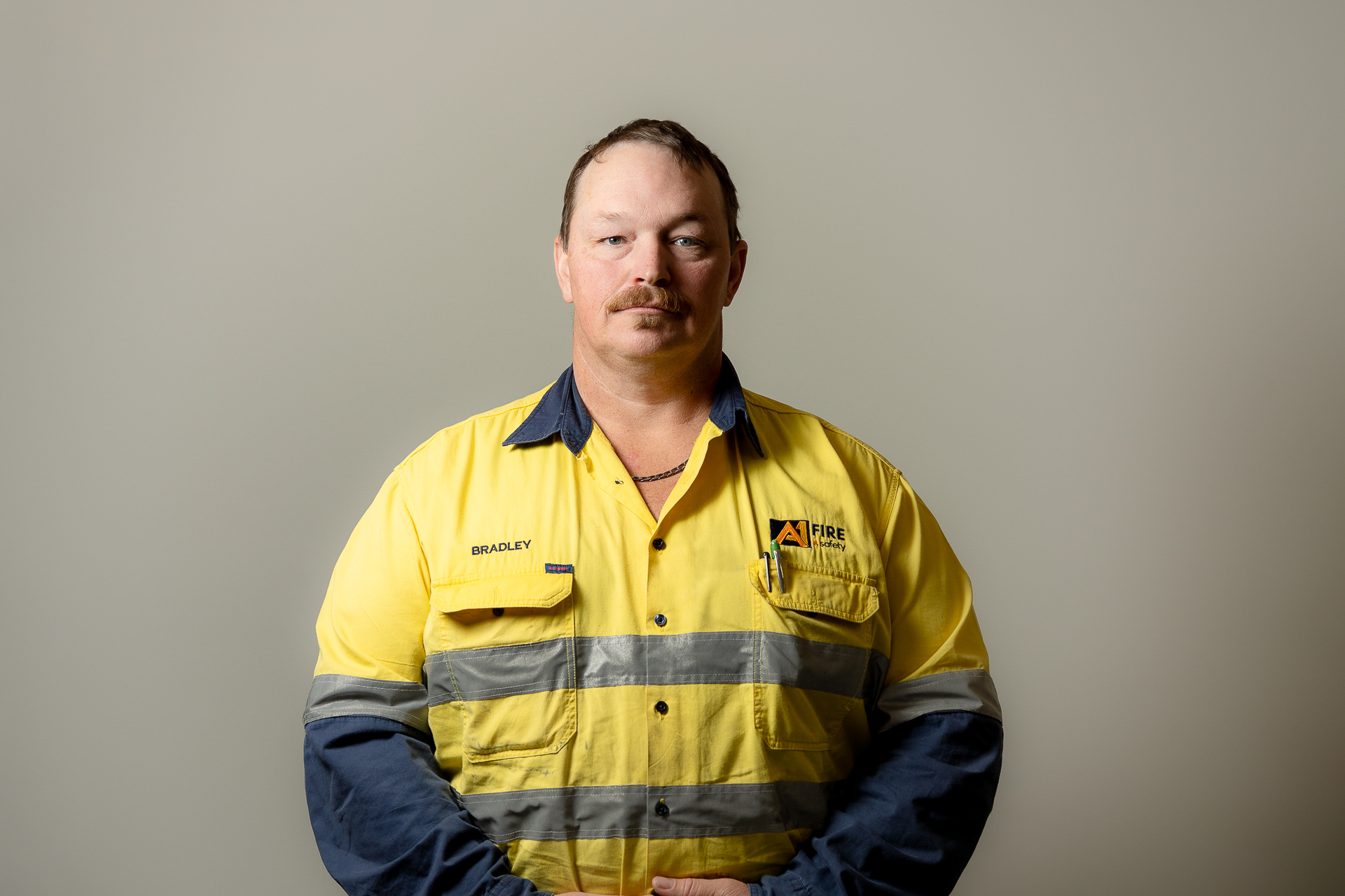 Man in a yellow high-visibility work shirt with reflective stripes, standing against a neutral background. The shirt has a logo and a name tag.