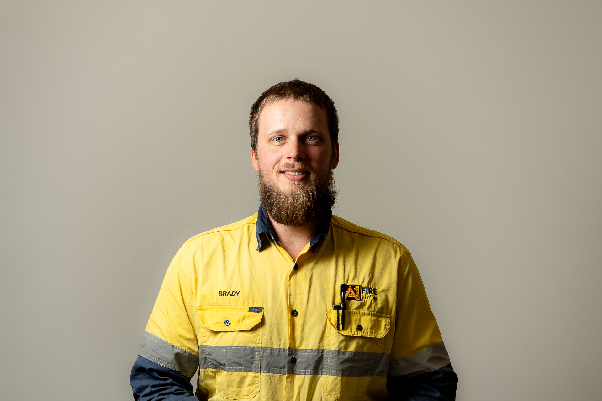 A man with a beard smiles while wearing a yellow high-visibility work shirt with blue accents and reflective stripes. The background is plain gray.