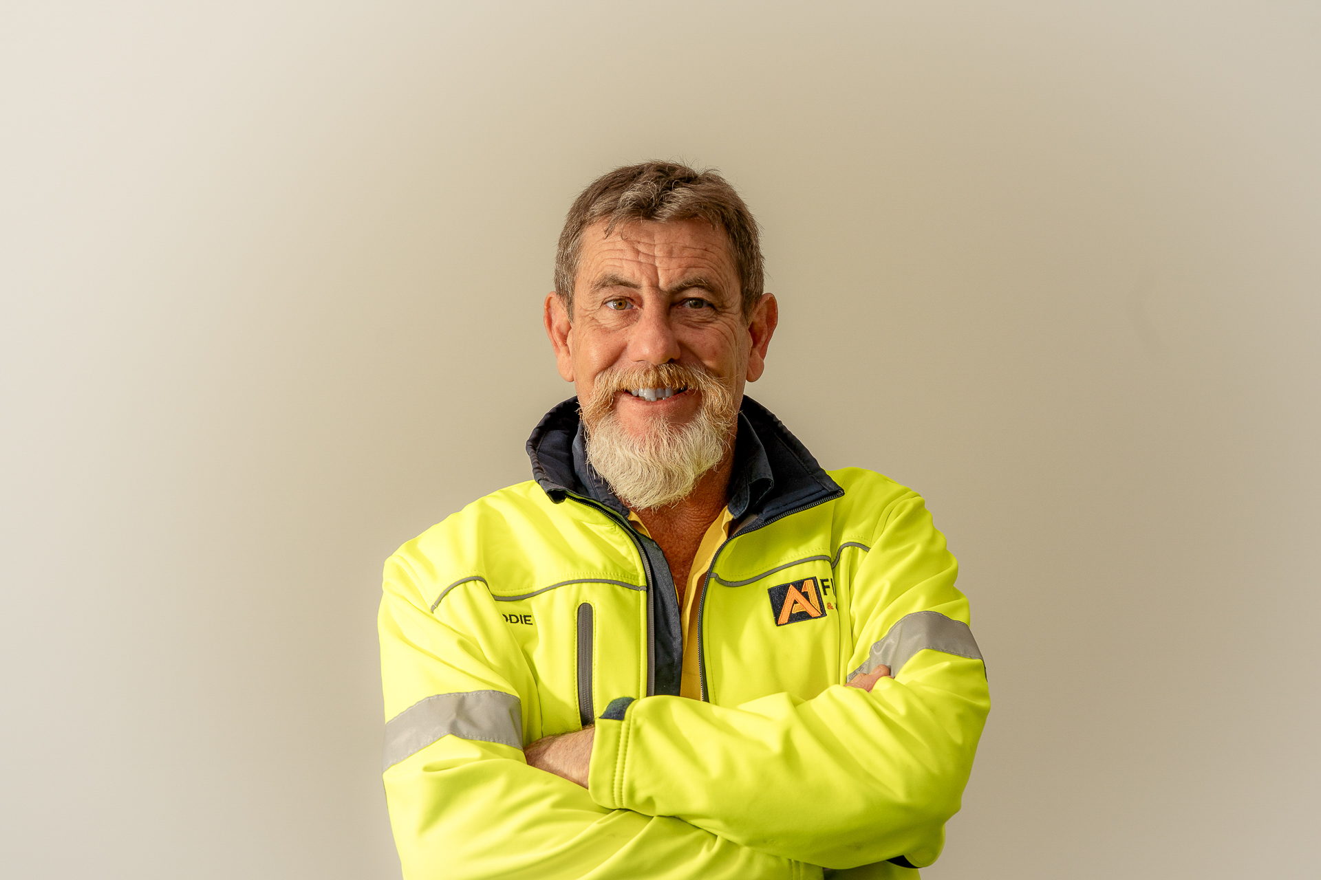 A man with a beard and short hair smiles warmly, wearing a bright yellow jacket with reflective stripes, standing against a plain light background.