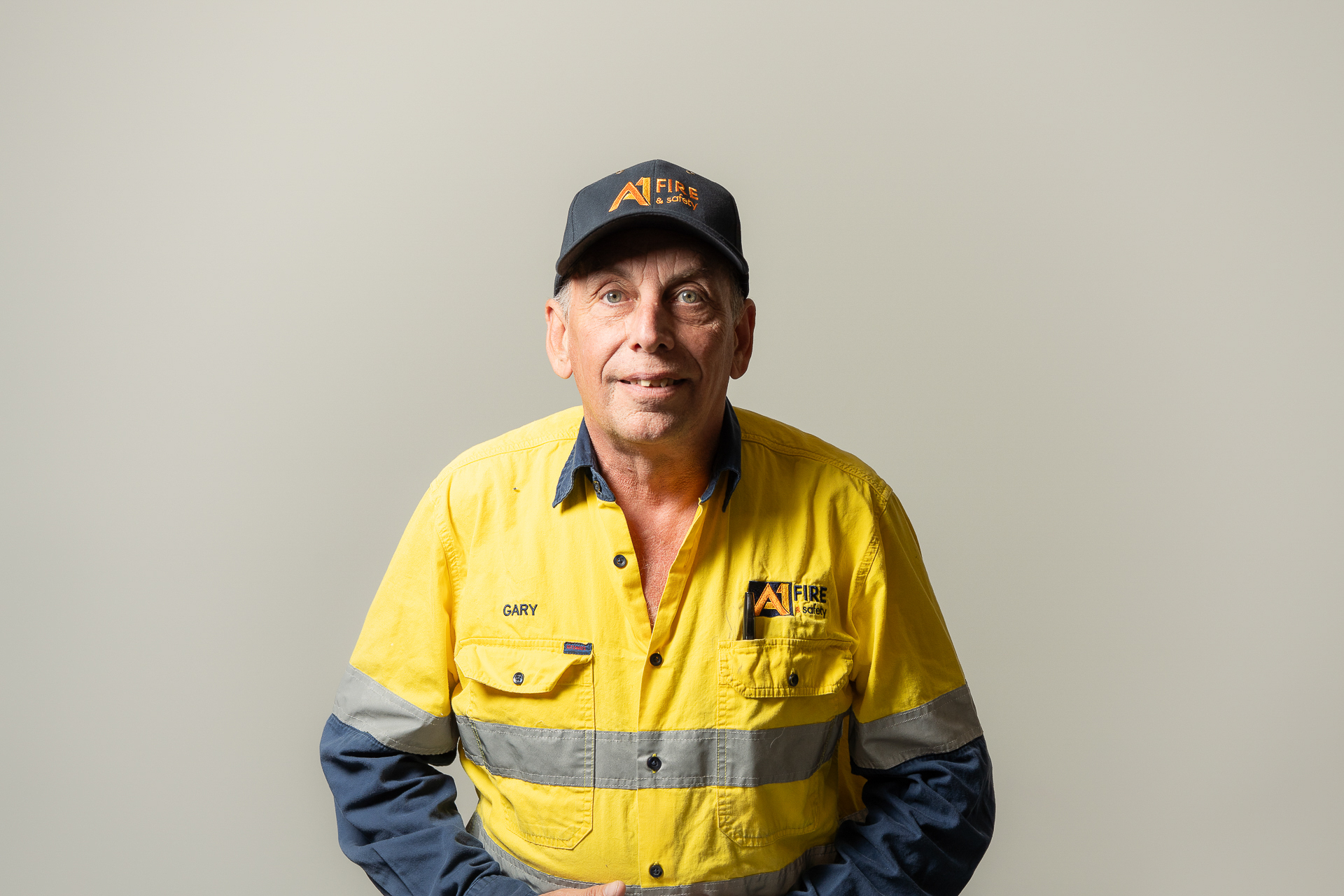 Man in a yellow high-visibility work shirt and cap with company logos stands smiling against a plain backdrop, exuding a friendly, professional vibe.