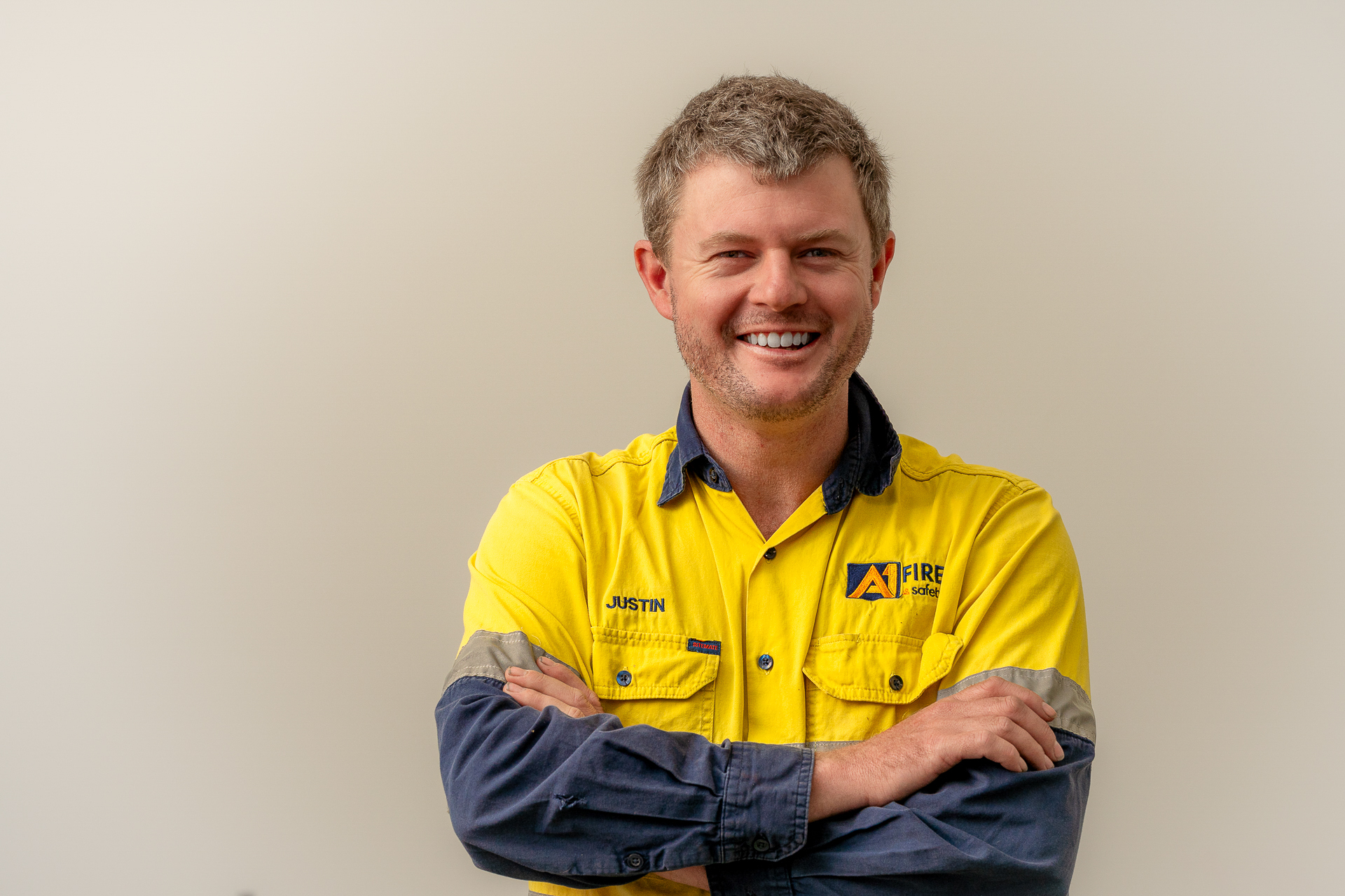A person in a yellow and blue uniform stands smiling with arms crossed. The background is a plain, light color, conveying a friendly and approachable tone.