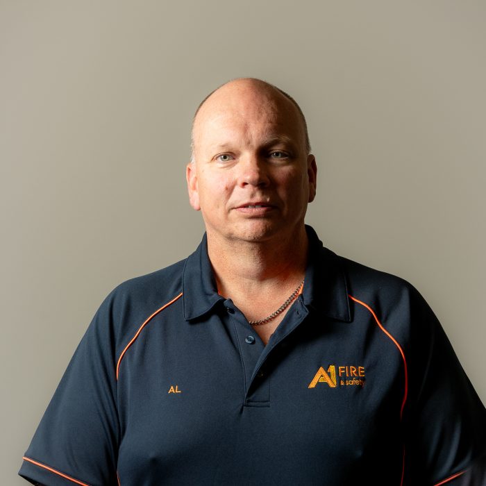 Bald man in a navy polo with "Al" and "A1 Fire" logos, stands against a plain gray background, looking confidently at the camera.