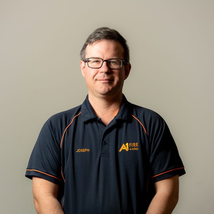 A man wearing glasses and a navy blue polo shirt with orange accents stands against a neutral background. The shirt features the name "Joseph" and a "Fire Academy" logo. He has a composed expression.
