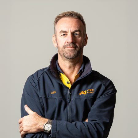 A person poses confidently with arms crossed, wearing a navy jacket over a yellow shirt. The jacket has "Aire" and "Fire & Safety" logos, set against a plain background.