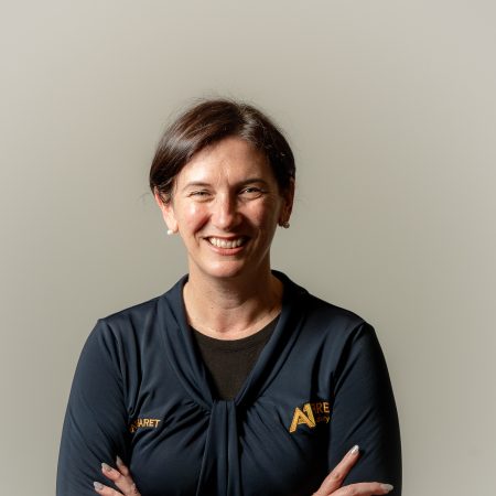 Smiling woman with dark hair, wearing a navy blue top, arms crossed, stands against a light gray background. Her expression conveys warmth and confidence.