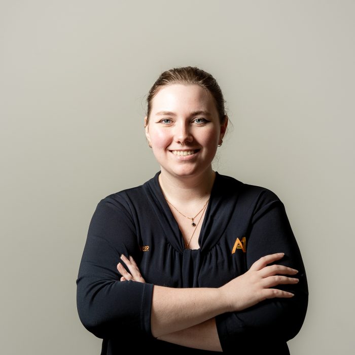 A smiling person stands confidently with arms crossed against a plain background, wearing a dark top with a subtle logo. The tone is professional and approachable.